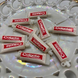 May include: A collection of nine miniature white toothpaste tubes with red "Colgate" lettering. The tubes have a silver cap and a small hole at the top. The tubes are arranged on a white and gold iridescent surface.