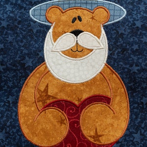 Könnte beinhalten: Ein brauner Teddybär, der einen rot-weiß-blauen Hut trägt und ein rotes Herz mit weißem Rand hält. Der Bär hat einen weißen Bart und einen weißen Schnurrbart. Der Hintergrund ist ein blauer Stoff mit weißen Sternen.