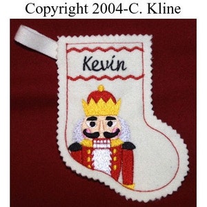 Puede incluir: Calcetín de fieltro blanco con un diseño de cascanueces bordado en rojo y blanco. El calcetín tiene un borde festoneado rojo y blanco y un lazo para colgar. El calcetín está personalizado con el nombre "Kevin".