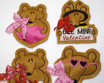 Könnte beinhalten: Vier Filz-Valentinstags-Anhänger mit braunen Bären in verschiedenen Designs. Ein Bär hält ein rosa Herz, ein anderer hat eine rosa Schleife, ein anderer hat eine rote Schleife und die Worte "XOXO" und der letzte Bär hat eine rosa Sonnenbrille und eine rosa Schleife.