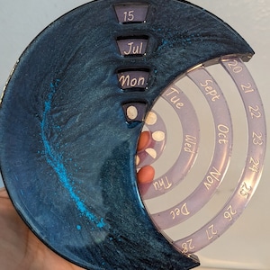 Pode incluir: Um calendário em forma de lua azul escuro e turquesa com uma sobreposição de plástico transparente. A sobreposição tem um design circular com os dias da semana e os meses do ano impressos nela. O calendário está configurado para 15 de julho.