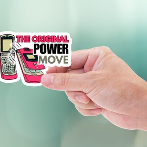 May include: A sticker featuring two retro flip phones in pink and silver. The sticker has the text "THE ORIGINAL POWER MOVE" in a bold font. The sticker is held by a hand against a blurred background.
