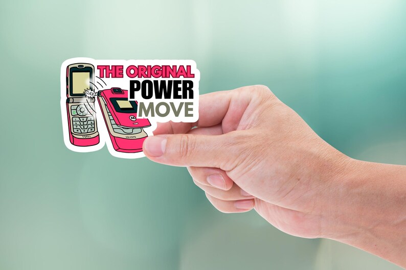 May include: A sticker featuring two retro flip phones in pink and silver. The sticker has the text "THE ORIGINAL POWER MOVE" in a bold font. The sticker is held by a hand against a blurred background.