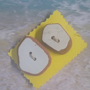May include: Two decorative buttons with a light brown edge and white center, each with a string for sewing. They rest on a yellow, scalloped-edge paper, set against a blurred background of ocean waves. These buttons are ideal for craft projects.