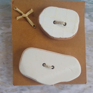 May include: A craft project featuring a brown cardboard base with two large, off-white, irregularly shaped buttons. The buttons have a textured surface and are attached with twine. A small, woven straw cross is affixed to the upper left corner.