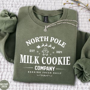 May include: A military green sweatshirt with the text "NORTH POLE MILK COOKIE COMPANY" in white. The design includes a gingerbread man, reindeer, and a sleigh. A silver necklace with a pendant is draped across the sweatshirt.