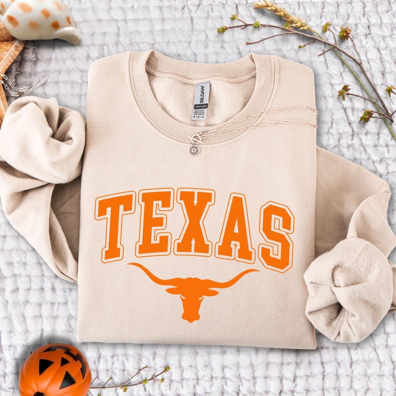 May include: A beige crewneck sweatshirt with the word "TEXAS" in orange block letters and a longhorn graphic. A silver necklace with a small pendant is draped across the sweatshirt. The sweatshirt is folded on a white textured surface.