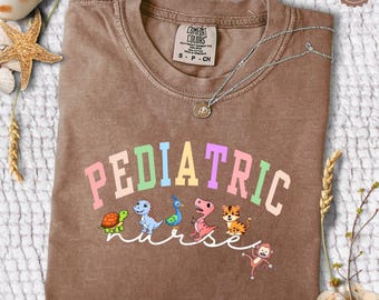 May include: A brown t-shirt with the text "PEDIATRIC nurse" in rainbow colors. The text is surrounded by cartoon animals including a turtle, dinosaur, peacock, T-Rex, tiger, and monkey.