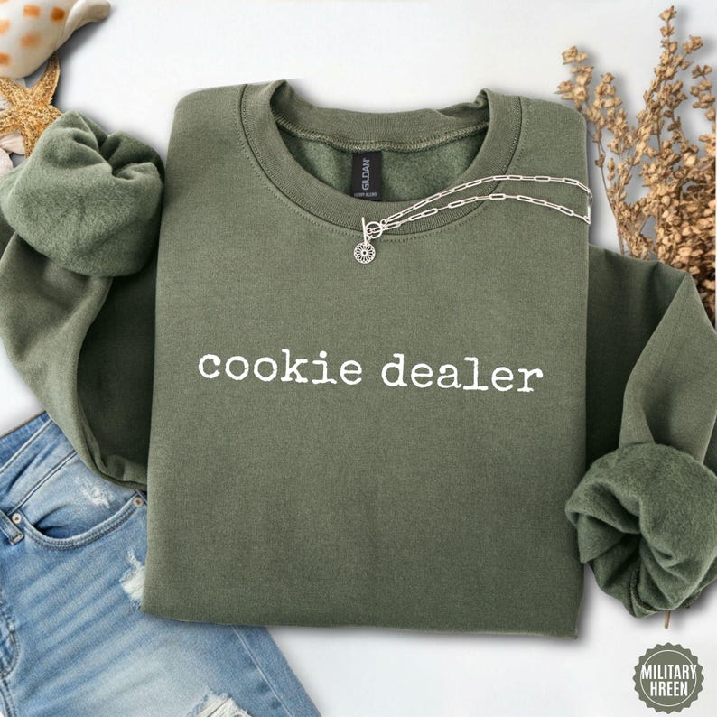 Puede incluir: Una sudadera verde oliva con el texto "cookie dealer" en blanco. Un collar plateado con un colgante circular est&aacute; drapeado en la parte delantera. La sudadera est&aacute; doblada, con las mangas y el dobladillo inferior visibles.