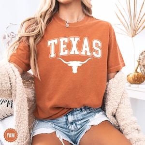 May include: Burnt orange t-shirt with the word "TEXAS" in white block letters and a white longhorn logo. The shirt has a crew neck and short sleeves. The shirt is made of a soft material.