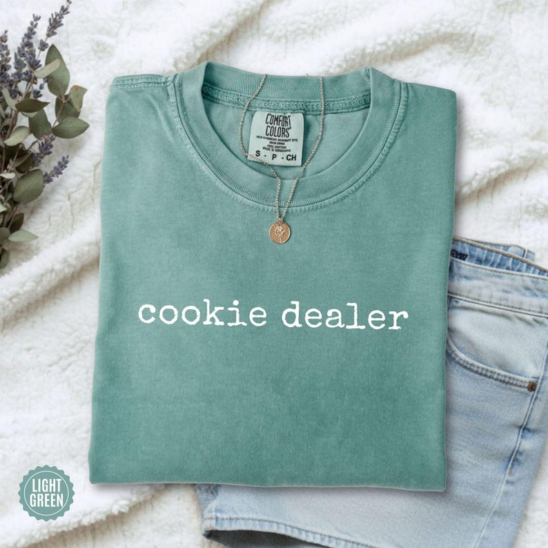 Puede incluir: Una camiseta verde claro con el texto "cookie dealer" en blanco. La camiseta est&aacute; doblada sobre una superficie blanca texturizada, con unos vaqueros azules visibles. Un collar dorado con un colgante est&aacute; sobre la camiseta.