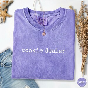 Puede incluir: Una camiseta violeta con la frase "cookie dealer" en blanco. La camiseta est&aacute; doblada y tiene cuello redondo. Un collar dorado con un colgante cuelga del cuello. La camiseta es de material suave.