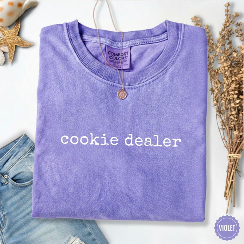 Puede incluir: Una camiseta violeta con la frase "cookie dealer" en blanco. La camiseta est&aacute; doblada y tiene cuello redondo. Un collar dorado con un colgante cuelga del cuello. La camiseta es de material suave.