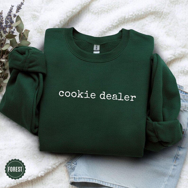 Puede incluir: Una sudadera verde oscuro con la frase "cookie dealer" en blanco en el pecho. La prenda est&aacute; doblada sobre una superficie blanca y texturizada. Una ramita de lavanda y unos vaqueros se ven en el fondo.