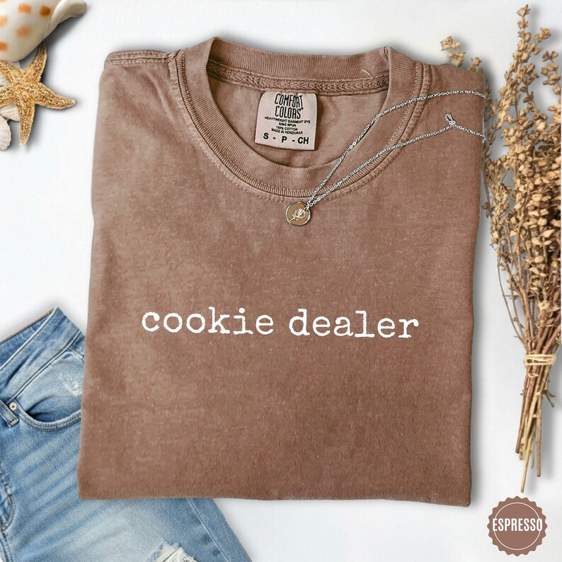 Puede incluir: Camiseta Comfort Colors color espresso con la frase "cookie dealer" en blanco. Un collar plateado con un peque&ntilde;o colgante descansa sobre la camiseta. La camiseta est&aacute; doblada y una peque&ntilde;a etiqueta es visible en el cuello.