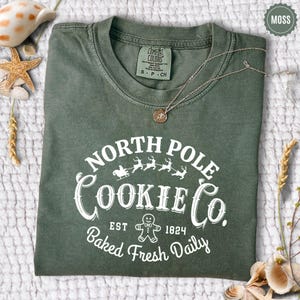 May include: A moss green t-shirt with the text "NORTH POLE COOKIE CO." in white. The design includes a gingerbread man, reindeer, and a sleigh. A silver necklace with a pendant rests on the shirt. The shirt is size S-P-CH.