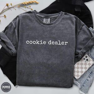 Puede incluir: Una sudadera gris oscuro con el texto "cookie dealer" en blanco. La sudadera est&aacute; doblada con unos vaqueros y una funda de tel&eacute;fono con un estampado de cuadros. Una ramita de trigo est&aacute; en el fondo.