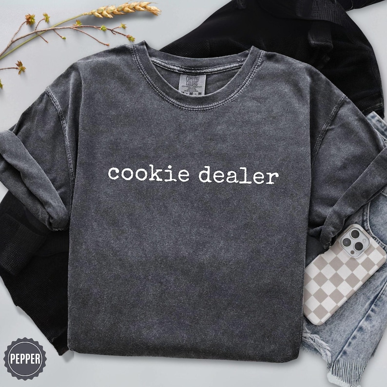 Puede incluir: Una sudadera gris oscuro con el texto "cookie dealer" en blanco. La sudadera est&aacute; doblada con unos vaqueros y una funda de tel&eacute;fono con un estampado de cuadros. Una ramita de trigo est&aacute; en el fondo.
