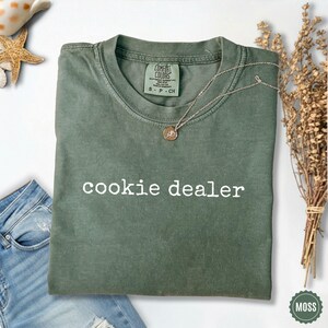 Puede incluir: Una camiseta verde musgo con el texto "cookie dealer" en blanco. Un collar plateado con un colgante descansa sobre la camiseta. La camiseta est&aacute; doblada y colocada sobre una superficie blanca con unos vaqueros.