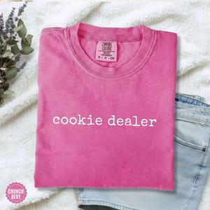 Puede incluir: Una camiseta rosa brillante con la frase "cookie dealer" en blanco. La camiseta, de la marca Comfort Colors, talla S-P-CH, est&aacute; doblada. Se encuentra sobre una superficie blanca texturizada, junto a unos pantalones cortos vaqueros azules.