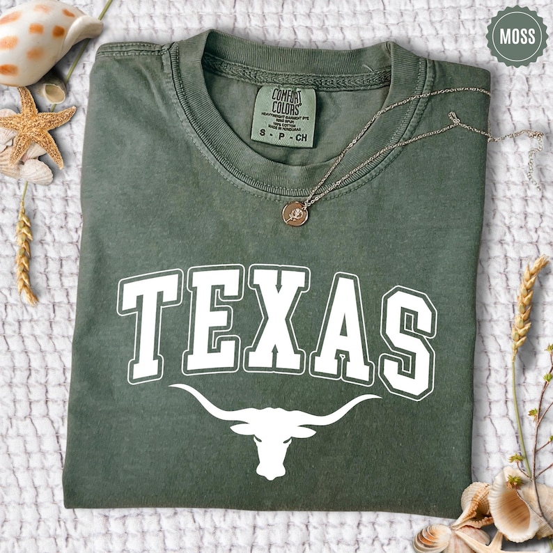 May include: A moss green t-shirt with the word "TEXAS" in white block letters and a white longhorn graphic. The shirt has a crew neck and a Comfort Colors tag. A silver necklace with a pendant rests on the shirt.
