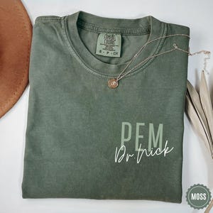 May include: A moss green Comfort Colors t-shirt with the text "PEM Dr. Trick" in white script. A delicate silver necklace with a small pendant rests on the shirt. A brown hat is visible in the upper left corner.