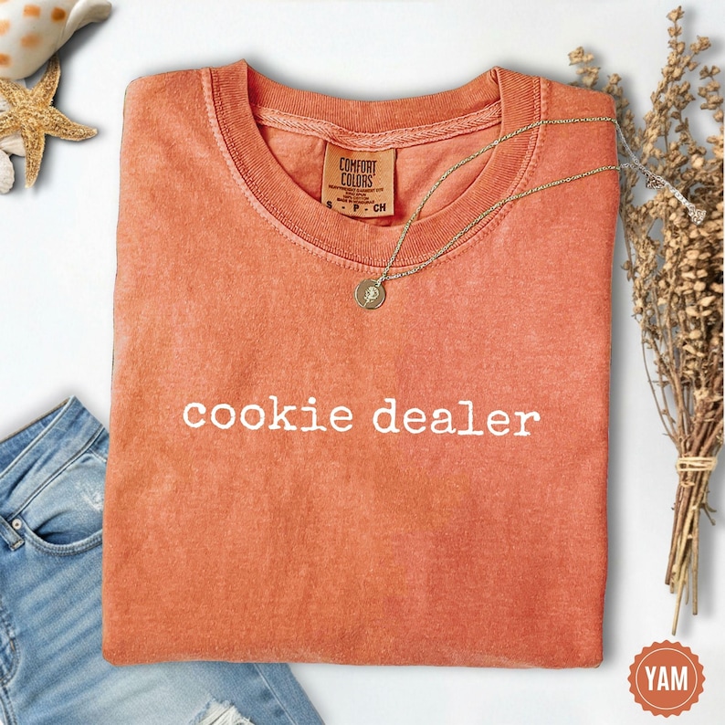 Puede incluir: Una camiseta de color coral con el texto "cookie dealer" en blanco. La camiseta tiene cuello redondo y mangas cortas. Un collar de oro con un colgante est&aacute; drapeado sobre la camiseta. La camiseta est&aacute; doblada y colocada sobre una superficie blanca.
