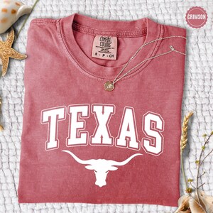 May include: Crimson-colored t-shirt with the word "TEXAS" in white block letters and a white longhorn graphic. The shirt has a crew neck and short sleeves. A silver necklace with a pendant rests on the shirt.