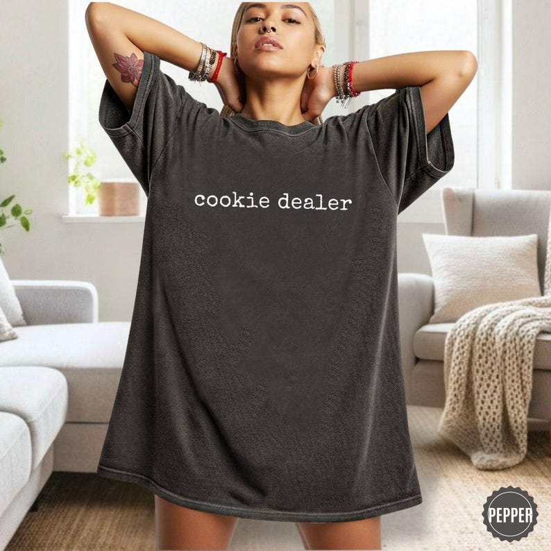 Puede incluir: Camiseta gris oscuro de gran tama&ntilde;o con el texto blanco "cookie dealer". La camiseta tiene un aspecto vintage ligeramente descolorido. La modelo lleva la camiseta con los brazos levantados.