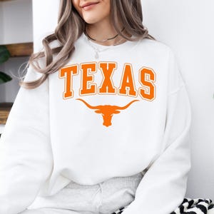 May include: White sweatshirt with the word "TEXAS" in orange block letters and a longhorn logo. The sweatshirt is a crew neck style and has a relaxed fit. The design is centered on the front of the shirt.