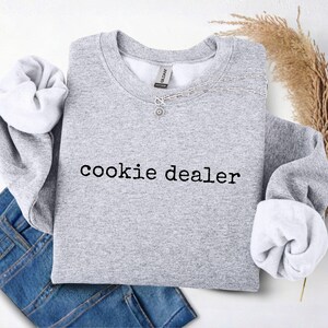 Puede incluir: Una sudadera gris jaspeada con la frase "cookie dealer" en negro. Un collar plateado con un colgante est&aacute; colocado en la parte delantera. La sudadera est&aacute; doblada sobre unos vaqueros azules.