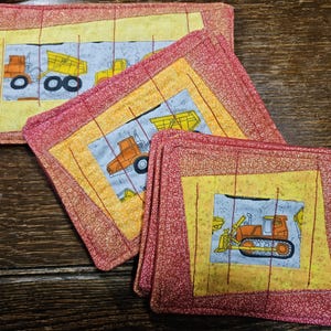 May include: Set of three quilted coasters featuring different construction vehicles. The coasters are red and yellow with a white background and feature a dump truck, a bulldozer, and a backhoe.
