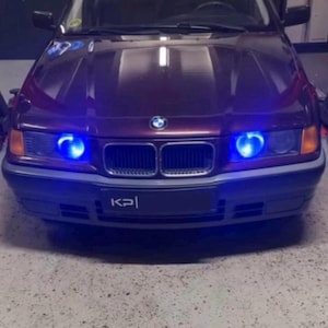 Op de afbeelding: Een donkerpaarse BMW met blauwe LED-koplampen en een "KPI" kentekenplaat. Een hand houdt een afstandsbediening met groene en rode knoppen.