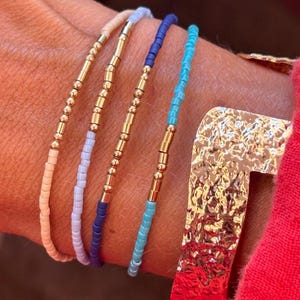 Op de afbeelding: Vier armbanden met kralen in pasteltinten: perzik, lavendel, marineblauw en turquoise. Elke armband heeft kleine, kleurrijke kralen afgewisseld met goudkleurige cilindrische kralen. De armbanden worden op een pols getoond, met een goudkleurige, getextureerde armband zichtbaar.
