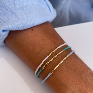 Puede incluir: Tres pulseras de cuentas blancas, turquesas y azul claro con detalles dorados. Las pulseras se llevan en un brazo, contra una camisa azul claro. Las pulseras están hechas de pequeñas cuentas.