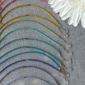 May include: A selection of colourful beaded necklaces in shades like red, pink, yellow, and blue. Each necklace has a delicate gold-toned clasp and extender chain. A white flower is in the background.