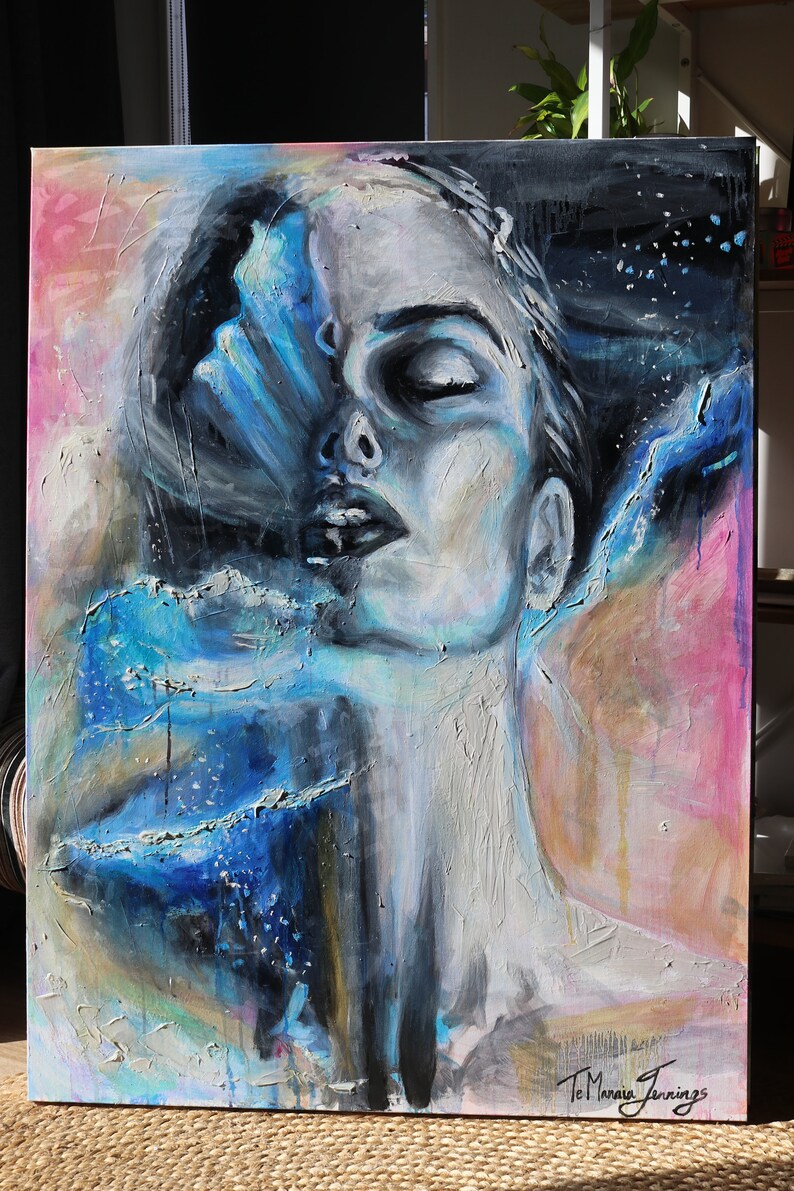May include: Abstract painting of a woman's face with blue and white paint, with a pink and yellow background. The painting is signed "Te Mania Jennings".