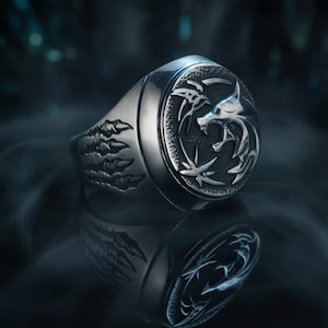 May include: A silver ring with a detailed wolf emblem on the face. The ring has a claw design on the side. The ring is set against a dark background with a smoky effect, highlighting the intricate details of the jewelry.