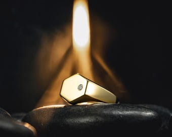 Puede incluir: Un anillo de sello dorado con una cara hexagonal y un pequeño diamante. El anillo está sobre un fondo oscuro con una luz suave y cálida que lo ilumina. El anillo descansa sobre una piedra negra.