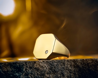 Puede incluir: Un anillo de sello dorado con una cara octogonal y un pequeño acento de diamante. El anillo está sobre una superficie oscura y texturizada, con un fondo dorado y cálido.