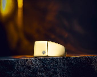 Puede incluir: Un anillo de sello dorado con una cara cuadrada y un pequeño diamante redondo. El anillo está sobre una superficie oscura y texturizada, con un fondo borroso y cálido.
