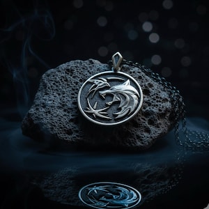 May include: A silver pendant necklace featuring a circular medallion with a wolf's head and other symbols. The pendant is displayed on a dark, textured rock, with a dark background. The necklace chain is also silver.