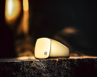 Puede incluir: Un anillo de sello dorado con una cara cuadrada y un pequeño diamante. El anillo se encuentra sobre un fondo oscuro y texturizado, con una cálida luz dorada.