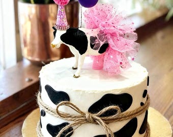 Könnte beinhalten: Ein runder Kuchen mit weißer Glasur und schwarzen Kuhflecken, umwickelt mit einer zu einer Schleife gebundenen Schnur. Obenauf steht eine Kuhfigur mit einem glitzernden rosa Partyhut mit Bommel, einem rosa Tüll-Tutu und kleinen rosa, weißen und lila Ballons.