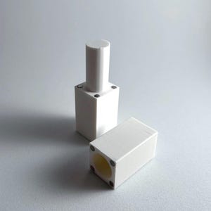 Op de afbeelding: Twee witte kunststof onderdelen met een cilindrische en een vierkante vorm. De onderdelen hebben een gat in het midden en zijn ontworpen om in elkaar te passen.