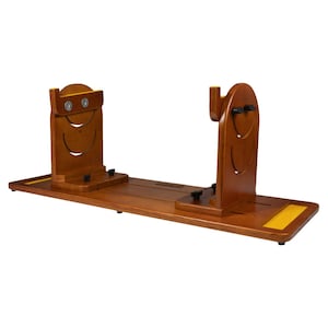 May include: A wooden jamón holder with a long, rectangular base and two upright supports. The supports have a curved design with a smiling face cutout. The holder is made of a warm, medium-toned wood and has a yellow accent on the end.