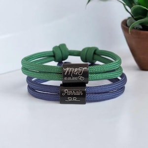 Puede incluir: Dos pulseras de cordón ajustables con cuentas rectangulares de metal oscuro. La pulsera verde superior está grabada con M&T, 10.03.2012 y dos corazones. La pulsera azul inferior tiene grabada la palabra Parrain y dos contornos de corazón.