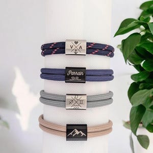 Peut inclure: Quatre bracelets en corde avec des fermoirs métalliques. Le premier affiche V, A, un cœur et une ancre. Le deuxième est noir avec le texte Parrain et deux cœurs. Le troisième présente quatre signes du zodiaque. Le dernier est noir avec un motif de montagne.