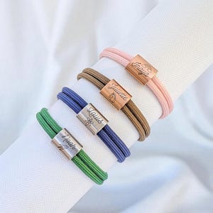 Peut inclure: Quatre bracelets personnalisés avec des bracelets en cordon tressé rose, marron, bleu et vert. Chaque bracelet comporte un fermoir en métal gravé d'un nom et d'un petit motif. Les noms sont "Fraya", "Juliette", "Louis" et "Noah".