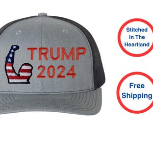 May include: Gray trucker hat with a black mesh back and a curved brim. The front features the text "TRUMP 2024" in red, with a flexed arm graphic in red, white, and blue.